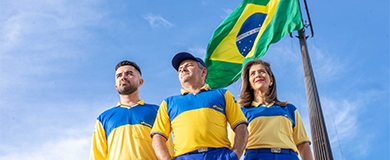Dois carteiros e uma carteira uniformizados em frente á um mastro com bandeira do Brasil. 